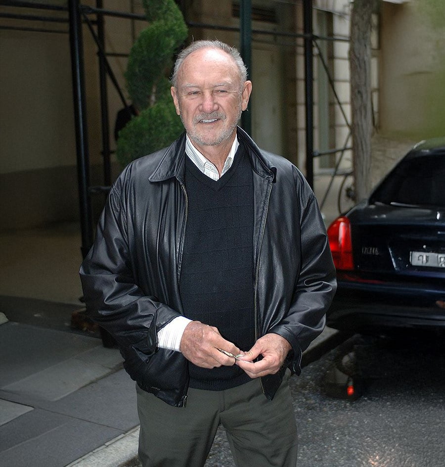 An older man with short gray hair and a beard is standing outdoors, wearing a black leather jacket over a black sweater and collared shirt. He is smiling and holding his glasses, with parked cars and greenery in the background.