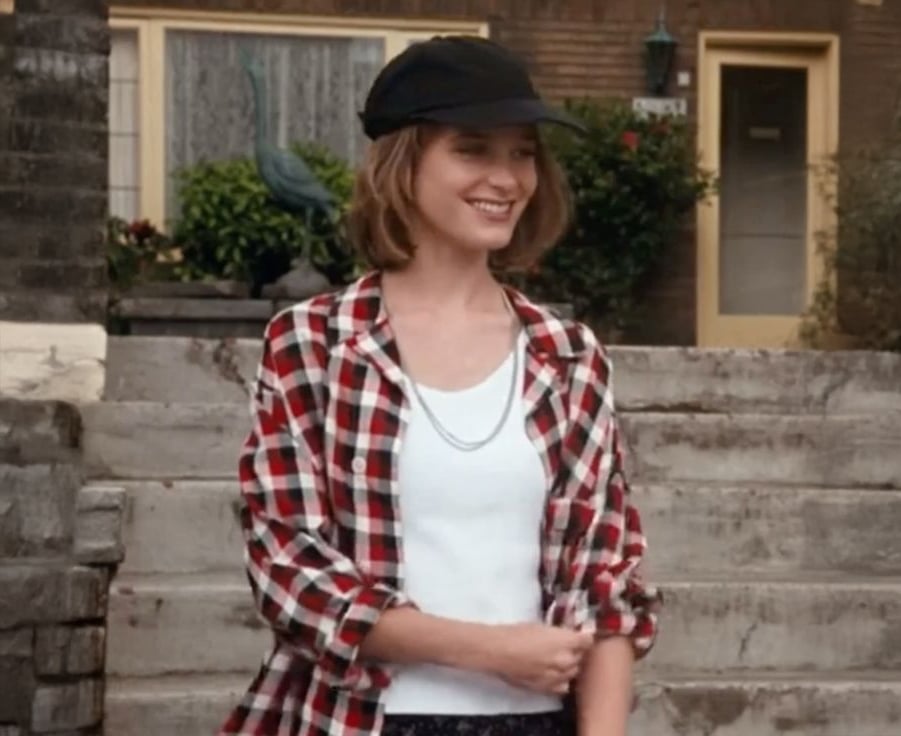 A woman wearing a black cap, red plaid shirt, and white top smiles while standing outside in front of steps leading to a house with yellow doors.