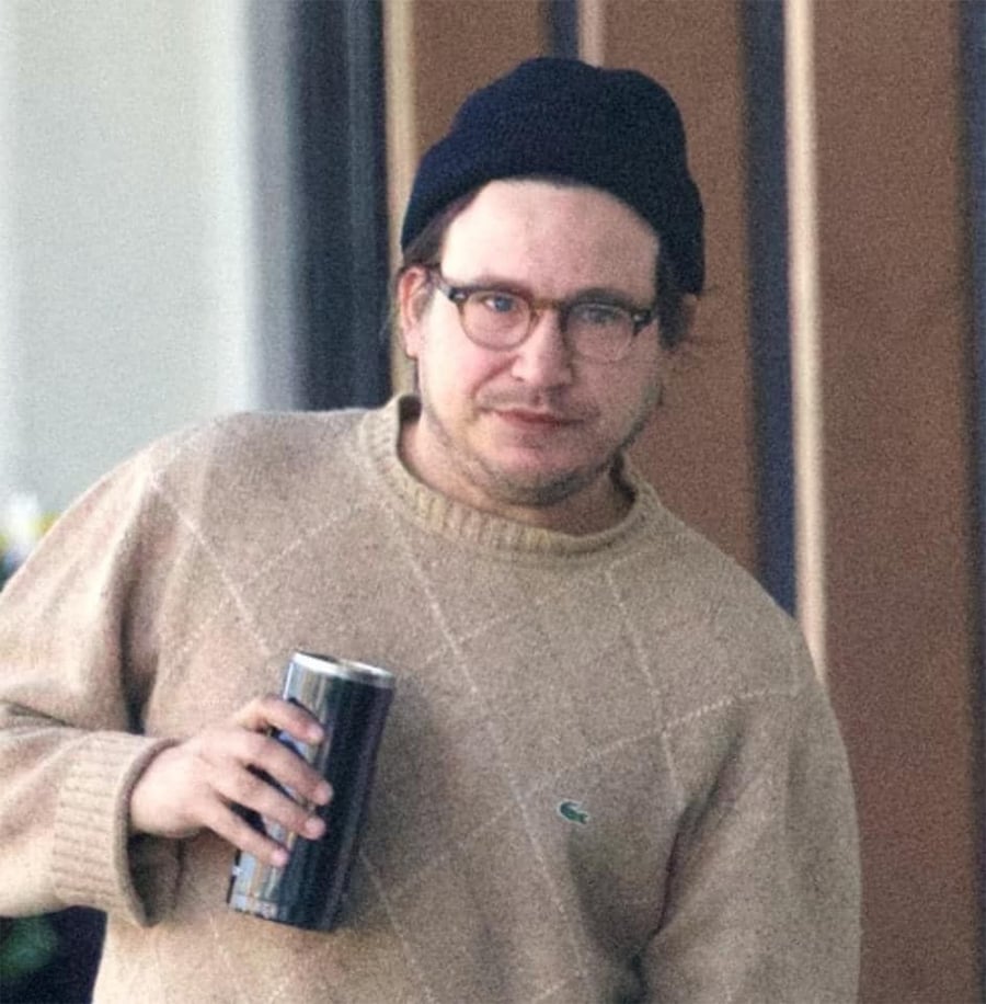 A person wearing glasses, a dark beanie, and a beige sweater with a crocodile logo holds a tumbler cup and looks toward the camera. Vertical panels are visible in the background.
