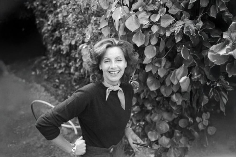 A woman with wavy hair smiles at the camera while standing next to a wall of leafy vines, wearing a sweater and scarf around her neck. A bicycle wheel is visible behind her.