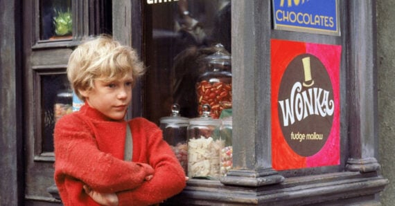 A young boy with blond hair and a red sweater stands outside a candy shop window filled with glass jars of sweets and signs reading "WONKA" and "CHOCOLATES." He looks thoughtfully to the side with arms crossed.