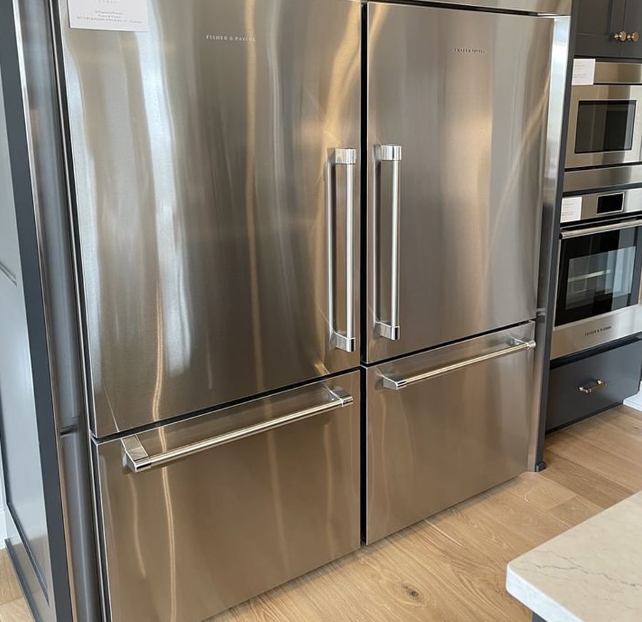 A modern stainless steel double-door refrigerator with two lower freezer drawers, set in a kitchen with built-in ovens and wood flooring.