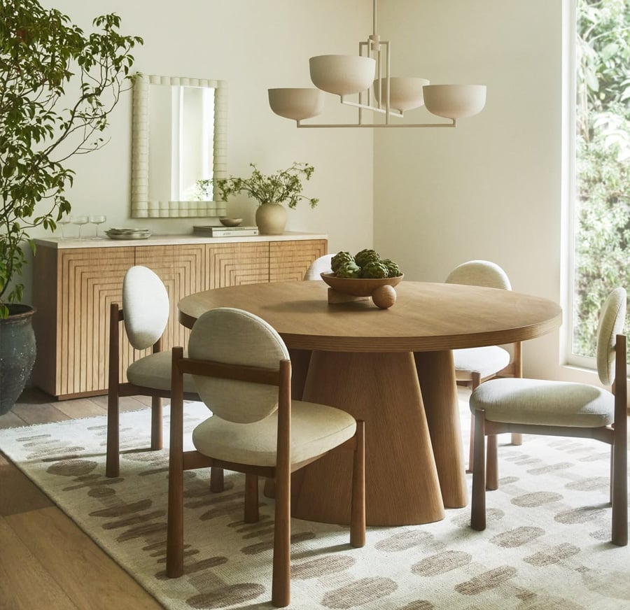 A modern dining room with a round wooden table, four upholstered chairs, a geometric chandelier, a patterned rug, and a sideboard with decor and a mirror. Large windows let in natural light, and there's a potted plant in the corner.