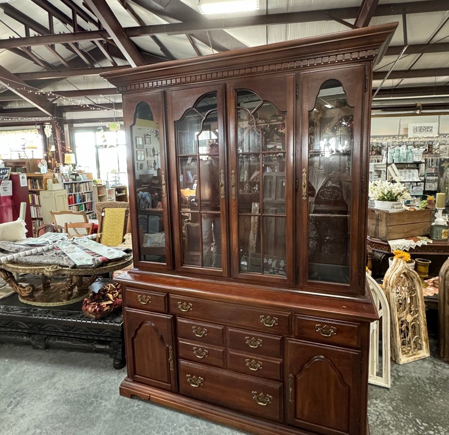 A large, dark wood china cabinet with glass doors on top and multiple drawers and cabinets below, displayed in a thrift or antique store with various other furniture and decor in the background.