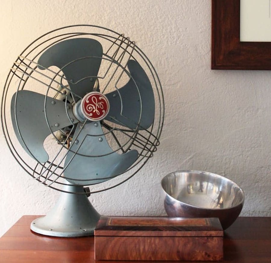 A vintage metal fan with a red GE logo sits on a wooden table next to a shiny metal bowl and a rectangular wooden box, against a light-colored wall.