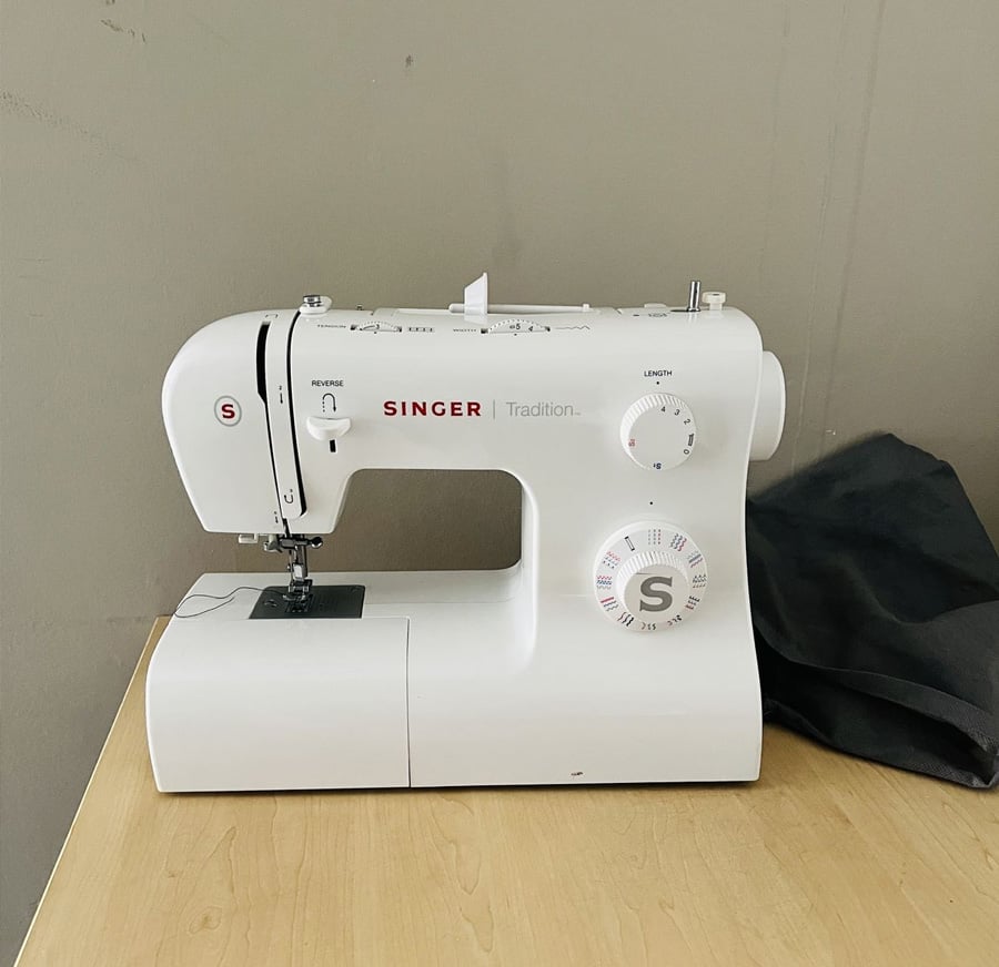 A white Singer Tradition sewing machine sits on a light wooden table against a plain gray wall, with a dark piece of fabric partially visible on the right.