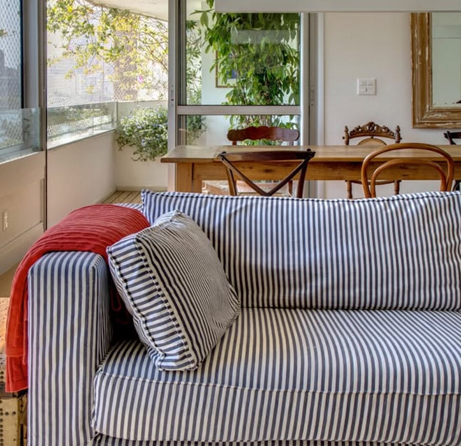 A blue and white striped sofa with matching cushions and a red throw sits in a bright room near a wooden dining table, with large windows and green plants outside.