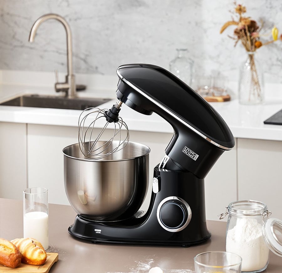 A black stand mixer with a stainless steel bowl and wire whisk sits on a kitchen counter, surrounded by a glass of milk, flour jar, sugar jar, and pastries. A modern kitchen sink and decor are visible in the background.