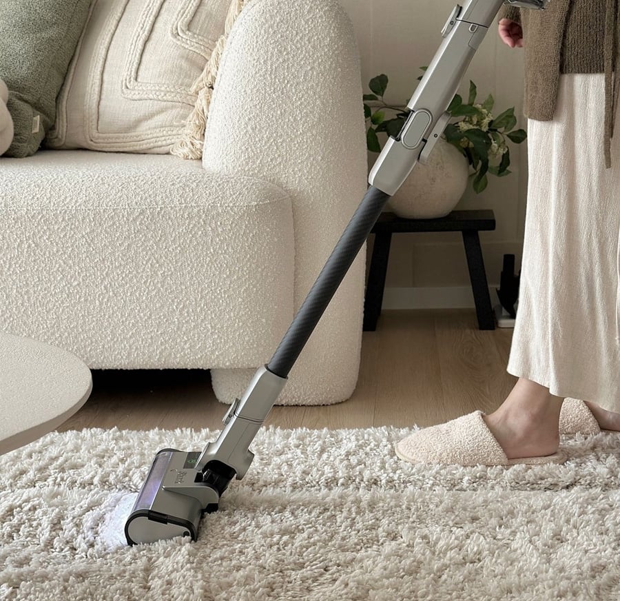 A person wearing slippers vacuums a plush, white rug in a cozy living room with a beige couch and decorative pillows in the background.
