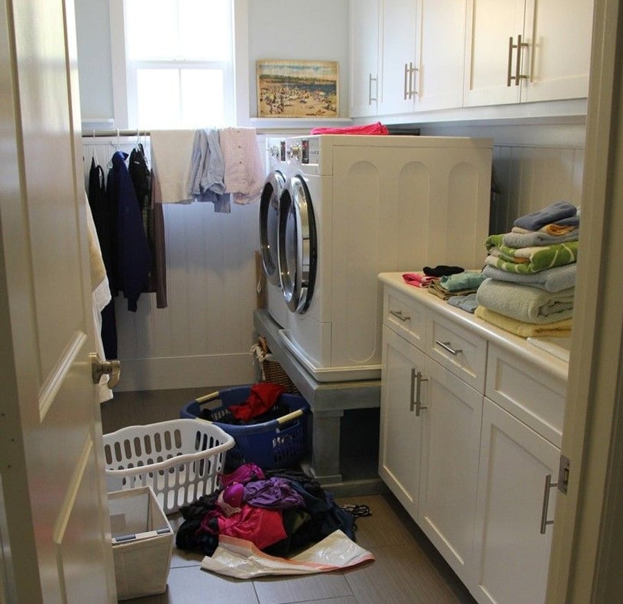 A laundry room with a washer and dryer, cabinets, baskets of laundry on the floor, folded towels on the counter, and clothes hanging to dry. A window lets in light above the machines.