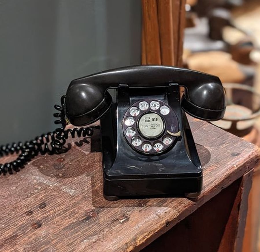A vintage black rotary dial telephone sits on a worn wooden surface, with a coiled cord connecting the handset to the base. The phone has metal details and visible numbers and letters on the dial.