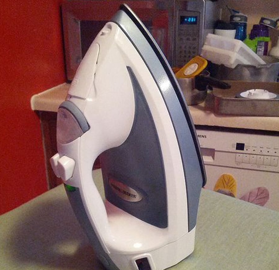 A household iron is standing upright on an ironing board in a kitchen, with appliances and dishes visible in the background.