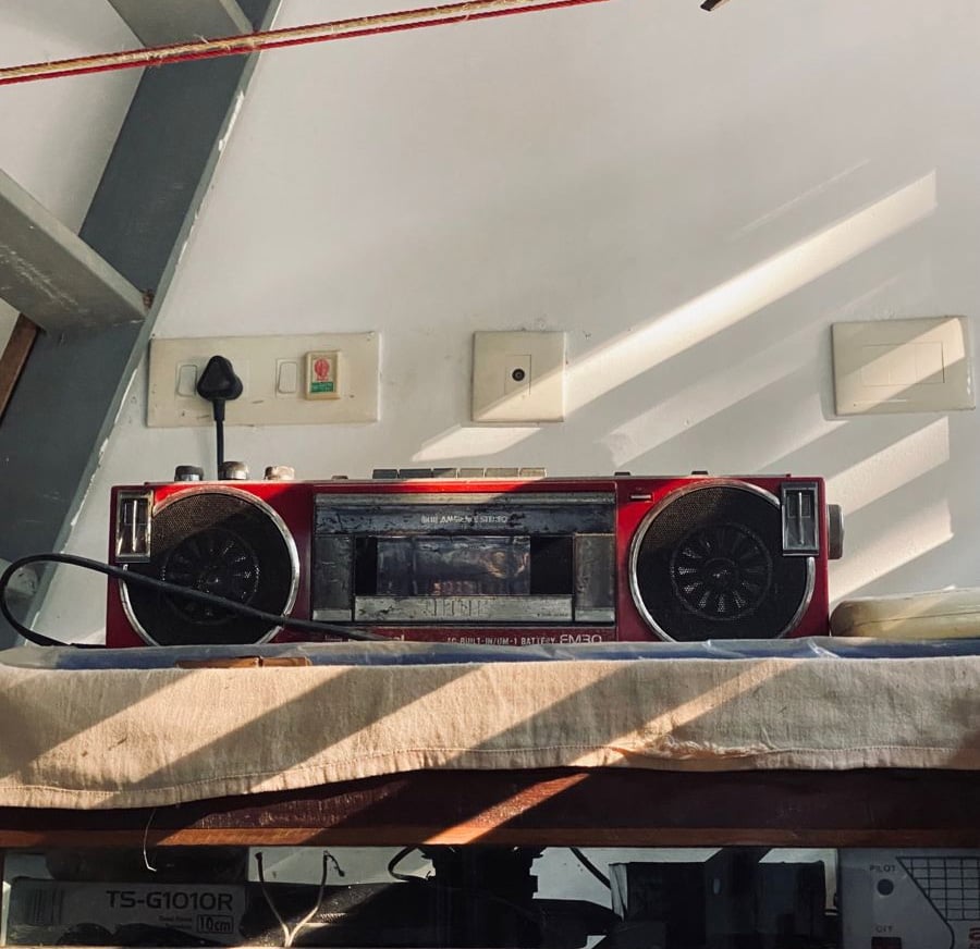 A red vintage boombox sits on a shelf against a wall with electrical outlets and sunlight casting shadows across the scene. A black power cord is plugged in above the boombox.