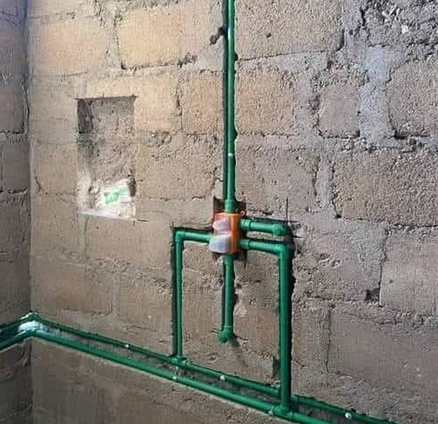 Green PVC pipes are attached to an unfinished concrete wall in a plumbing setup, with a plastic snack wrapper used as a makeshift connector at the intersection of the pipes.