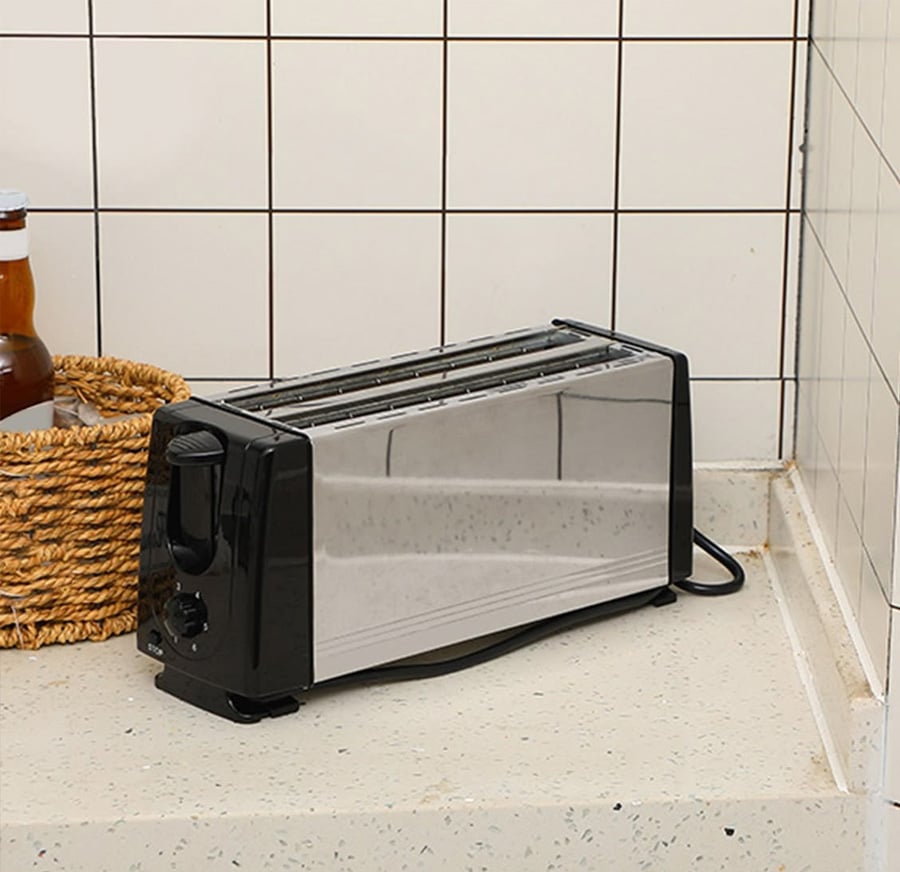 A stainless steel toaster with black accents sits on a speckled kitchen countertop near a basket and a bottle, with white tiled walls in the background.