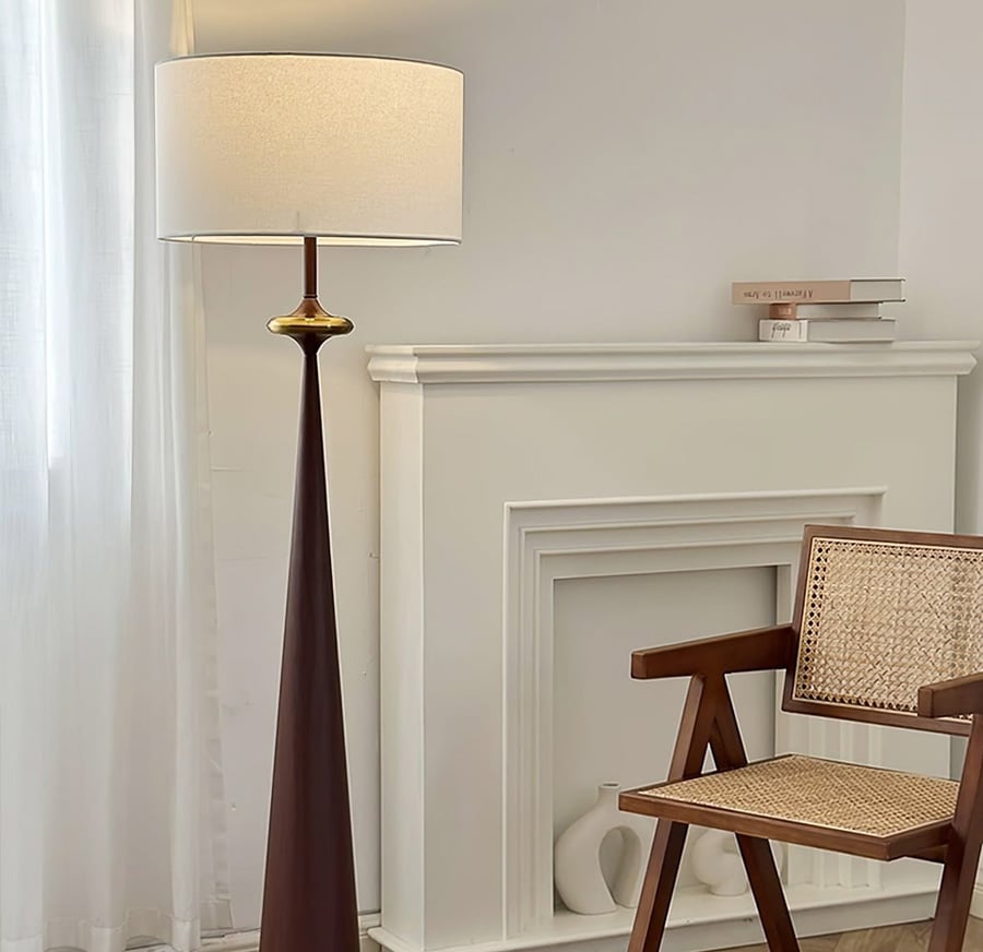A modern living room corner with a tall floor lamp, a wooden chair with a woven seat, a white fireplace mantel, books on top, and minimal decor, softly lit by natural light from a window with sheer curtains.