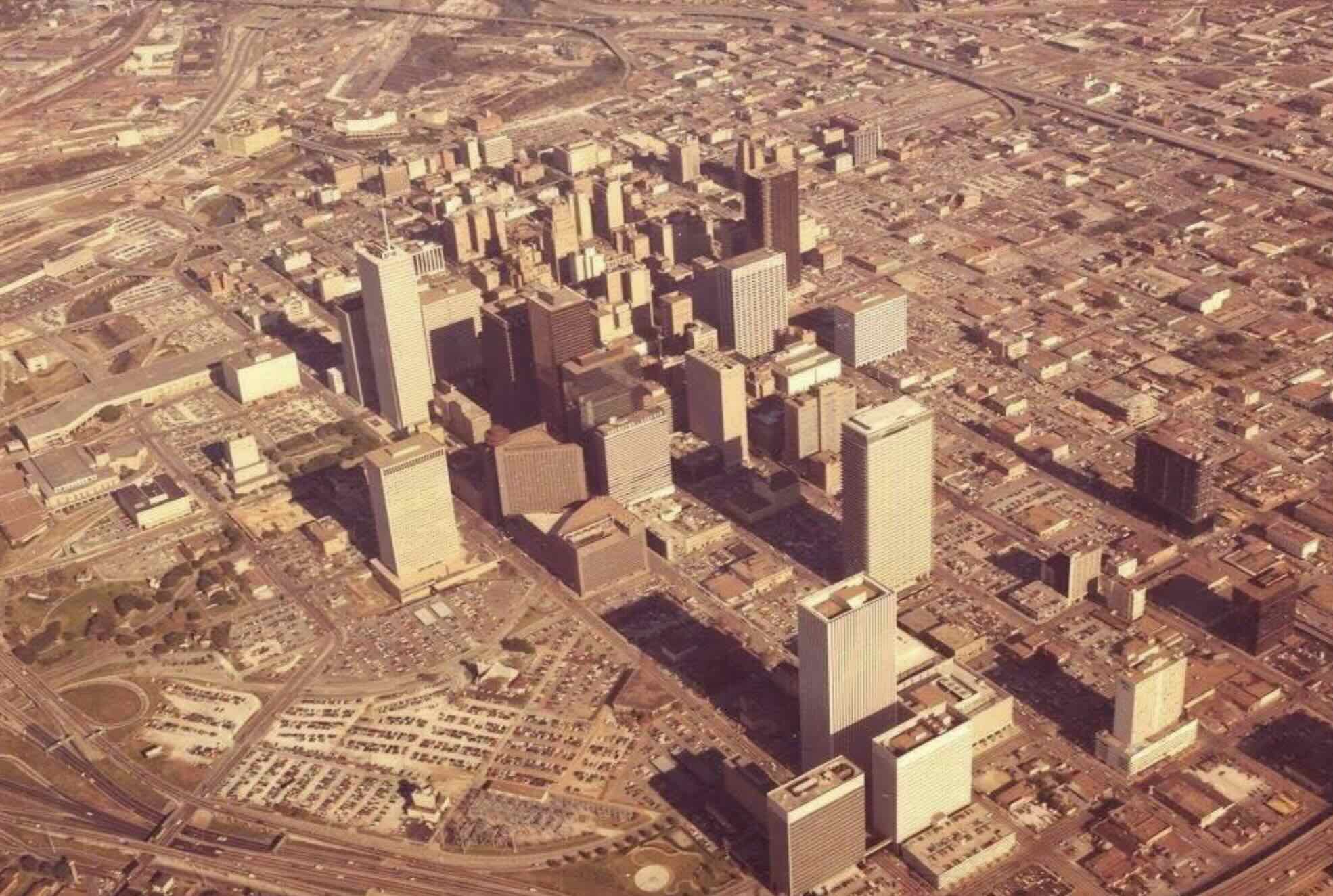 Aerial view of a city downtown with tall skyscrapers, grid-like streets, and numerous parking lots, likely taken in the 1970s based on the image color and car styles.
