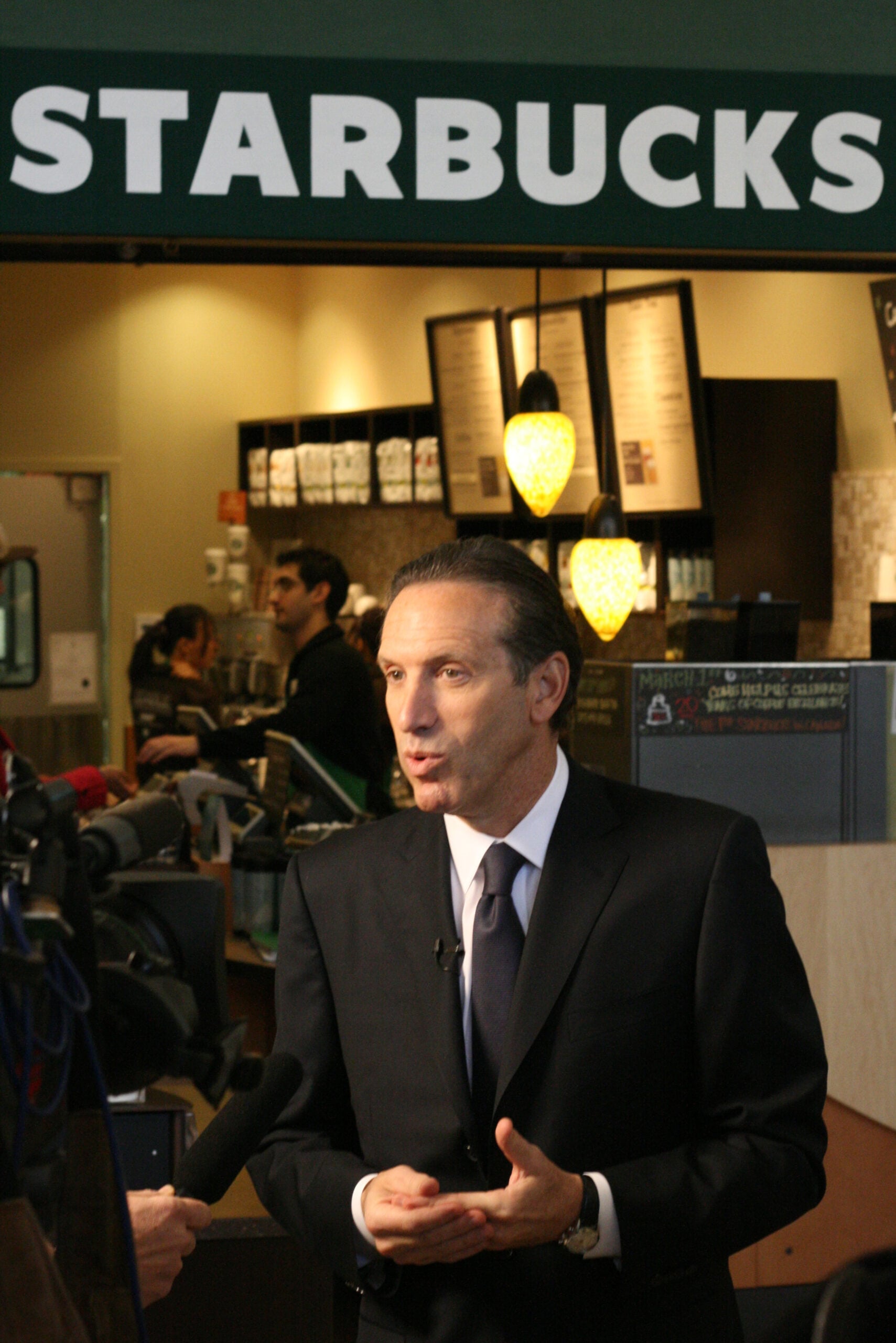 A man in a suit speaks to reporters inside a Starbucks coffee shop, standing under a large Starbucks sign, with staff and menu boards visible in the background.