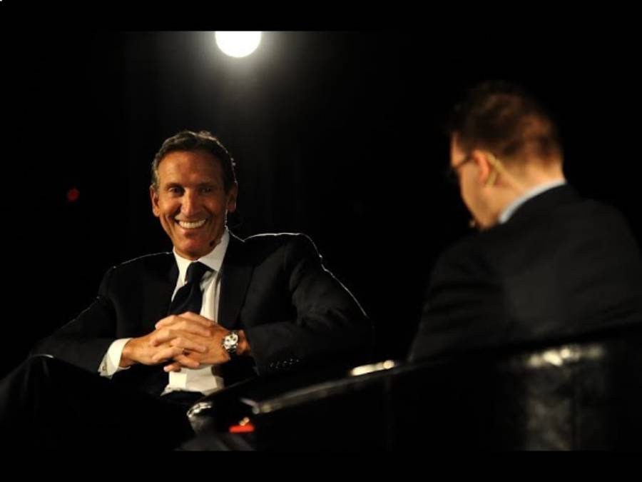 Two men in suits sit facing each other on a dimly lit stage, one smiling broadly under a spotlight, while the other has his back to the camera.