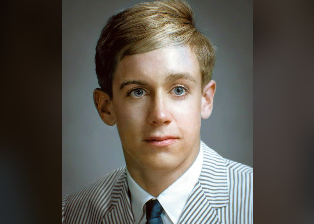 A young man with light skin, blond hair, and blue eyes wears a pale striped suit jacket, white shirt, and tie, posing against a neutral background.
