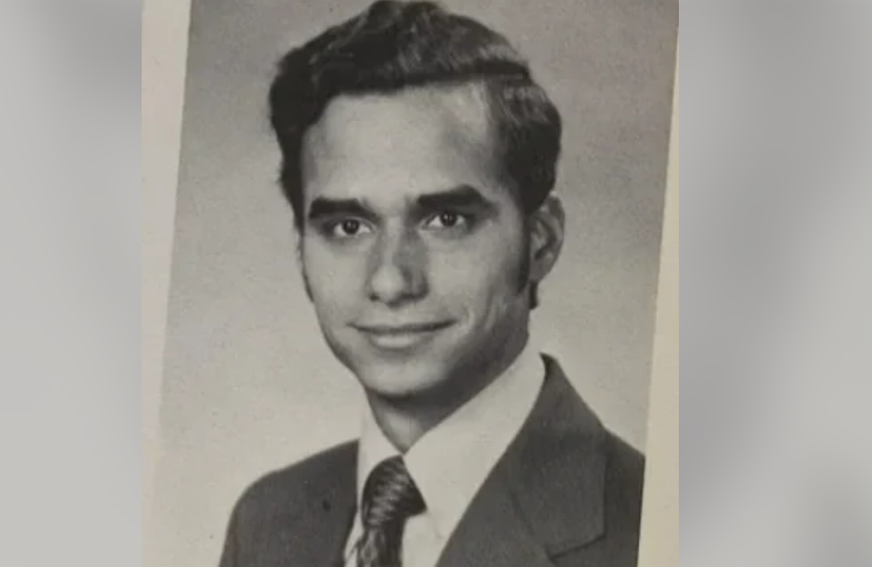 Black and white portrait of a young man in a suit and tie, with dark, neatly styled hair, looking at the camera and smiling slightly. The background is plain and the photo has a classic vintage appearance.
