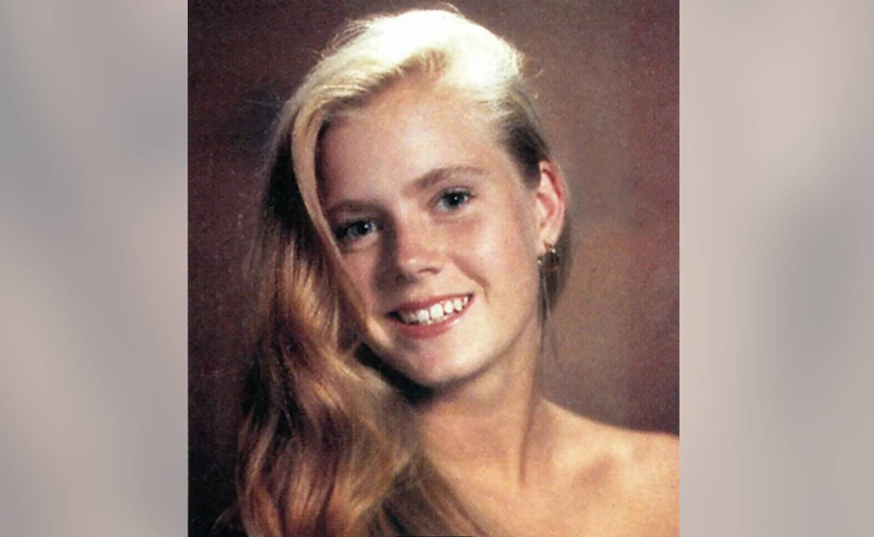 A young woman with long, wavy blonde hair smiles at the camera. She is wearing a small hoop earring and has bare shoulders against a plain, soft brown background.