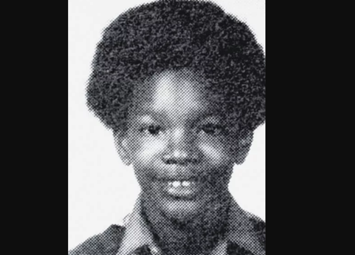 Black and white yearbook-style photo of a young boy smiling, with a round face, wide eyes, and an afro hairstyle, wearing a collared shirt. The image appears to be from the 1970s or 1980s.