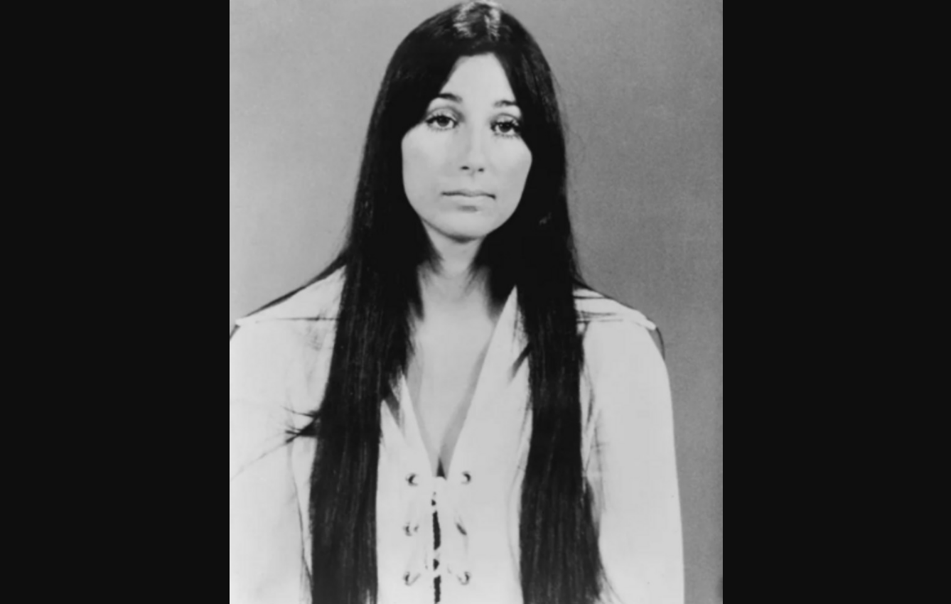 A black-and-white portrait of a woman with long, straight dark hair wearing a light-colored, lace-up top, facing the camera with a neutral expression against a plain background.