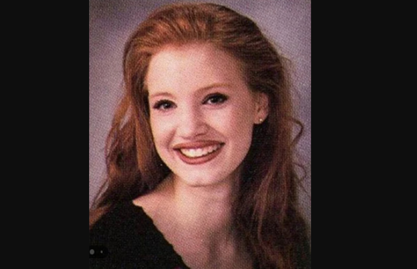 A young woman with long, wavy red hair is smiling at the camera. She is wearing a black top and has a fair complexion, with a neutral gray background behind her.