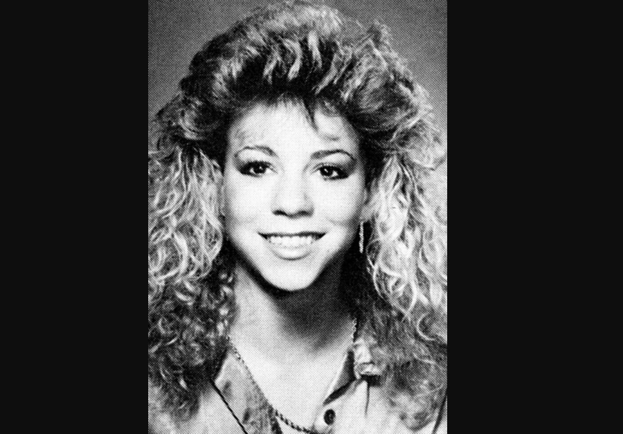Black and white portrait of a young woman with voluminous curly hair, smiling at the camera. She is wearing a collared shirt and a necklace. The background is plain and the photo has a slight vintage feel.