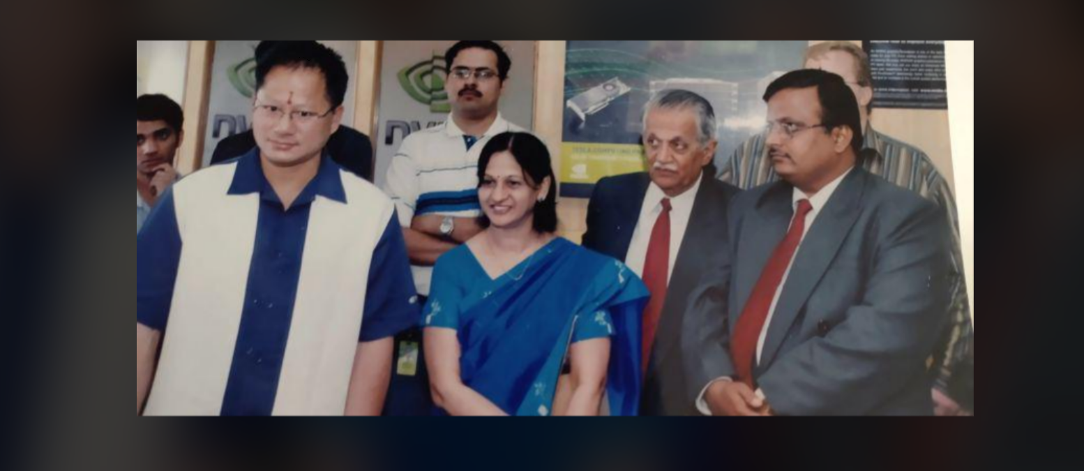 A group of six adults, including one woman in a blue saree and five men in formal wear, stand together indoors in front of posters, posing for a photo.