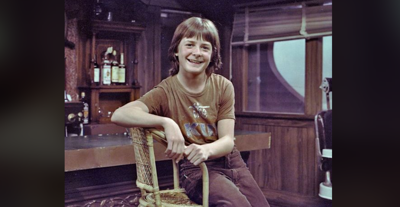 A smiling young boy with brown hair sits casually on a wicker chair in a cozy, wood-paneled room with bottles on a shelf and a window in the background. He wears a brown T-shirt and brown pants.