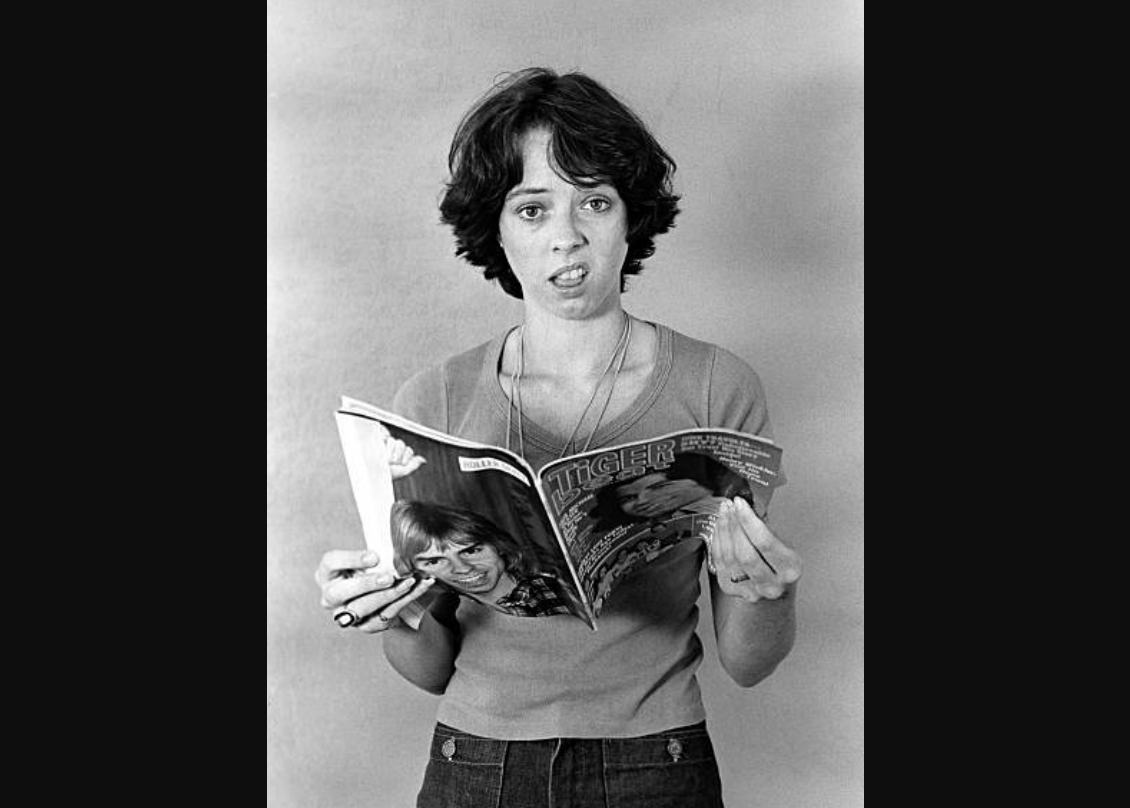 A person with short hair and a neutral expression holds an open magazine titled "TIGER" and looks slightly puzzled or confused, standing against a plain background.