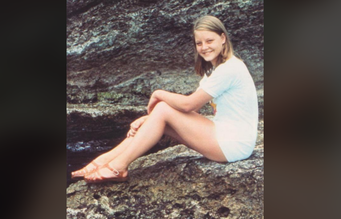 A young girl with blonde hair, wearing a white t-shirt and shorts, sits on a rocky surface outdoors. She is smiling and looking at the camera, with her arms resting on her knees.