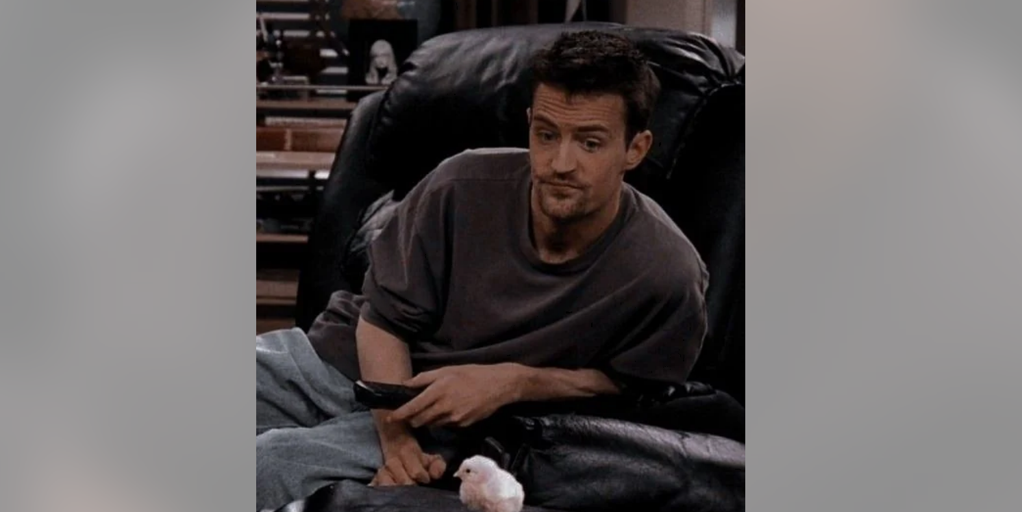 A man in a loose gray shirt sits on a black leather chair, holding a TV remote and looking down thoughtfully at a small white chick on his lap.