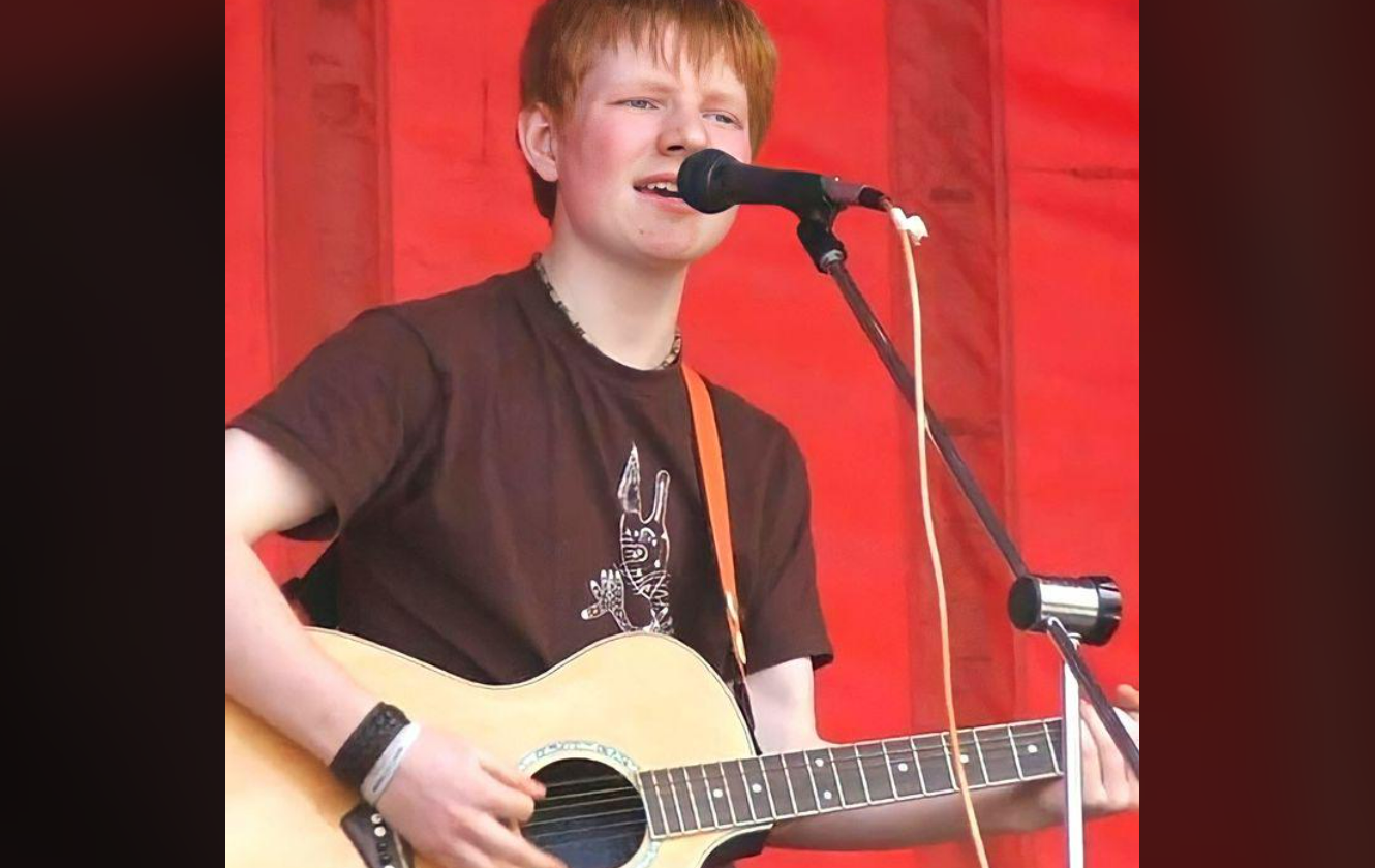 A young person with light skin and red hair sings into a microphone while playing an acoustic guitar on stage, with a bright red background behind them.