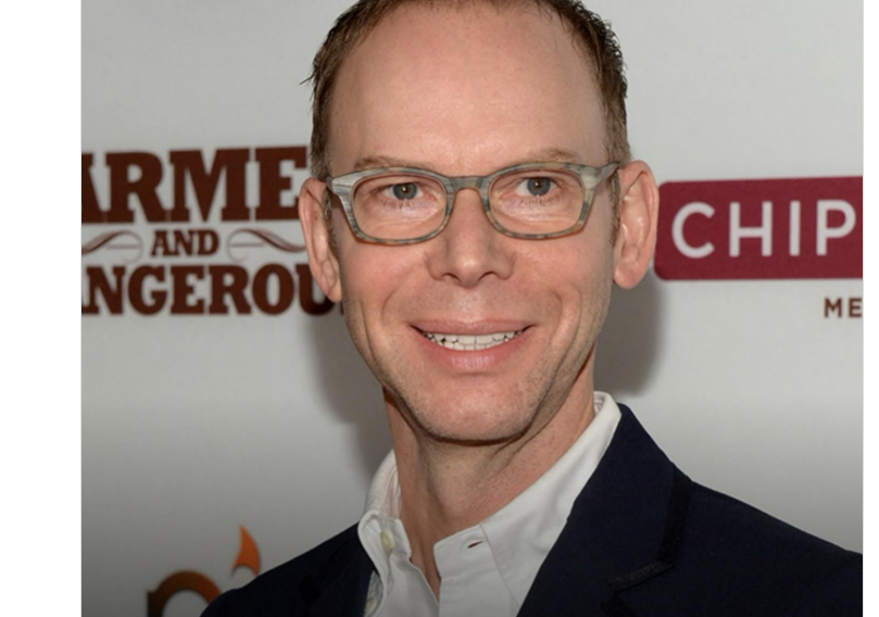 A man with short light brown hair and glasses smiles at the camera, wearing a dark blazer over a white shirt. The background features partial logos and text on a white backdrop.