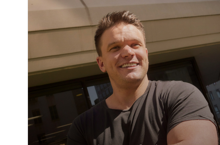 A smiling man with short hair stands outside in front of a building, wearing a dark t-shirt with his arms crossed. Sunlight casts shadows on his face and the building behind him.