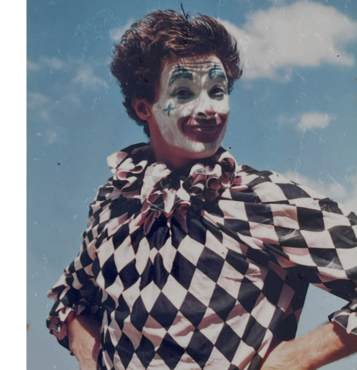 A person dressed as a clown with white face paint, exaggerated red lips, and blue designs poses with hands on hips, wearing a black and white diamond-patterned costume against a blue sky with clouds.