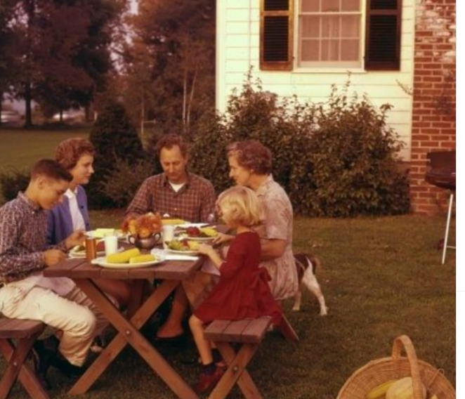 A family of five sits at a wooden picnic table outdoors, enjoying a meal together. There is a basket on the grass nearby, and a small dog stands beside the table. A house and shrubs are visible in the background.