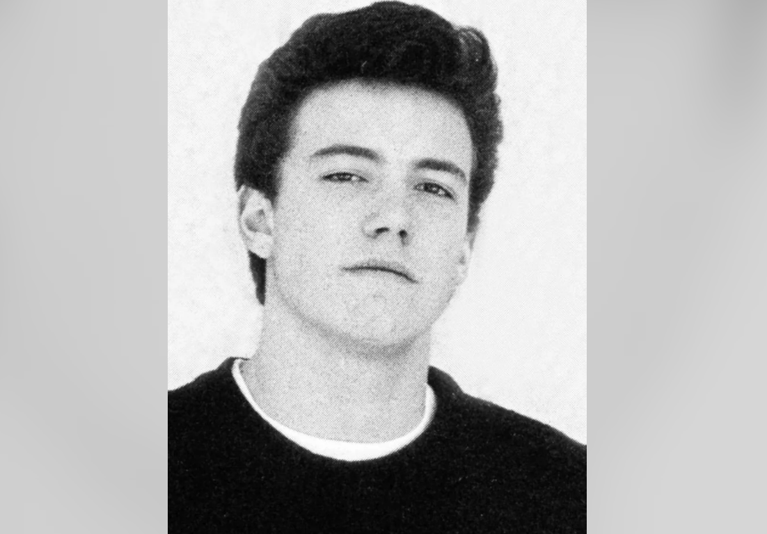 A young man with dark hair styled upward looks at the camera with a neutral expression. He is wearing a dark shirt over a light undershirt. The background is plain and light-colored.