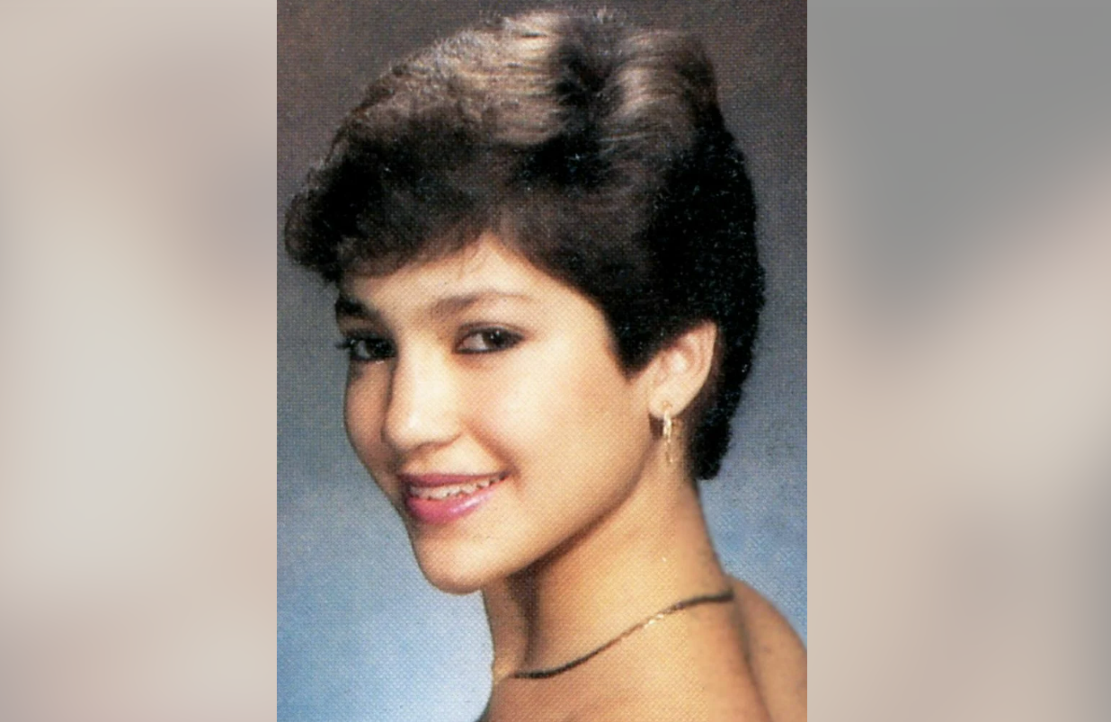 A young woman with short brown hair smiles at the camera, wearing a simple necklace and hoop earrings against a softly blurred, neutral background.