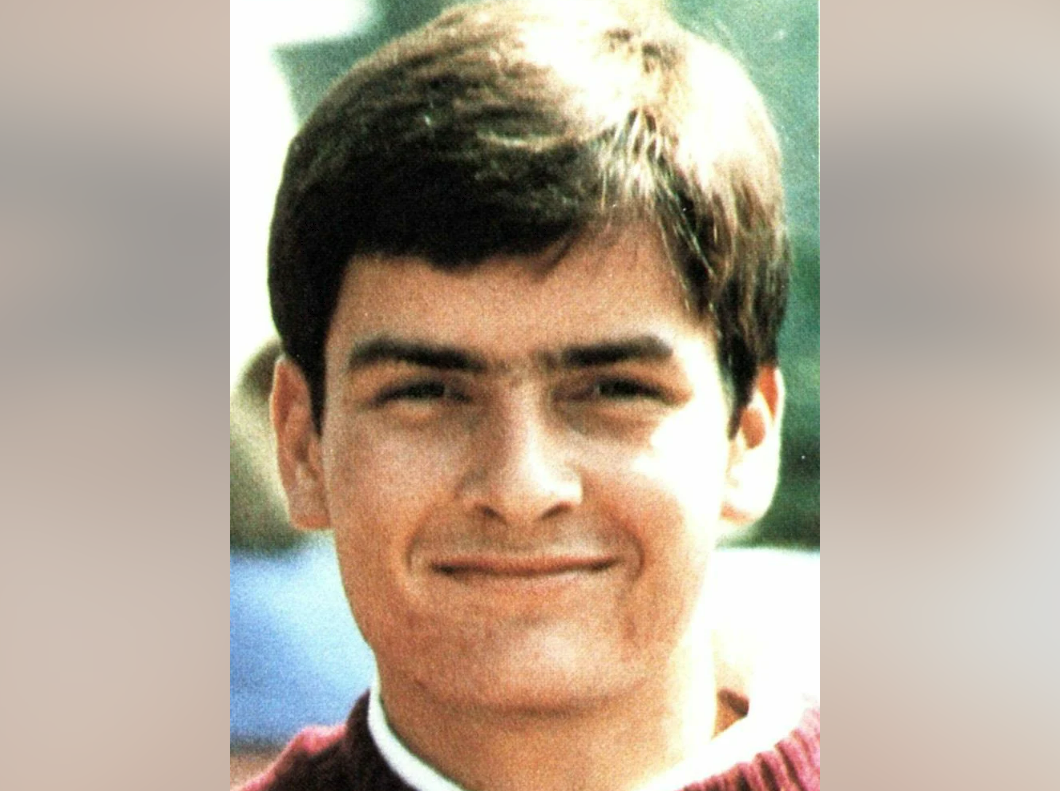 A young man with short dark hair smiles softly at the camera, wearing a maroon sweater over a white collared shirt. The background is blurred and outdoors.