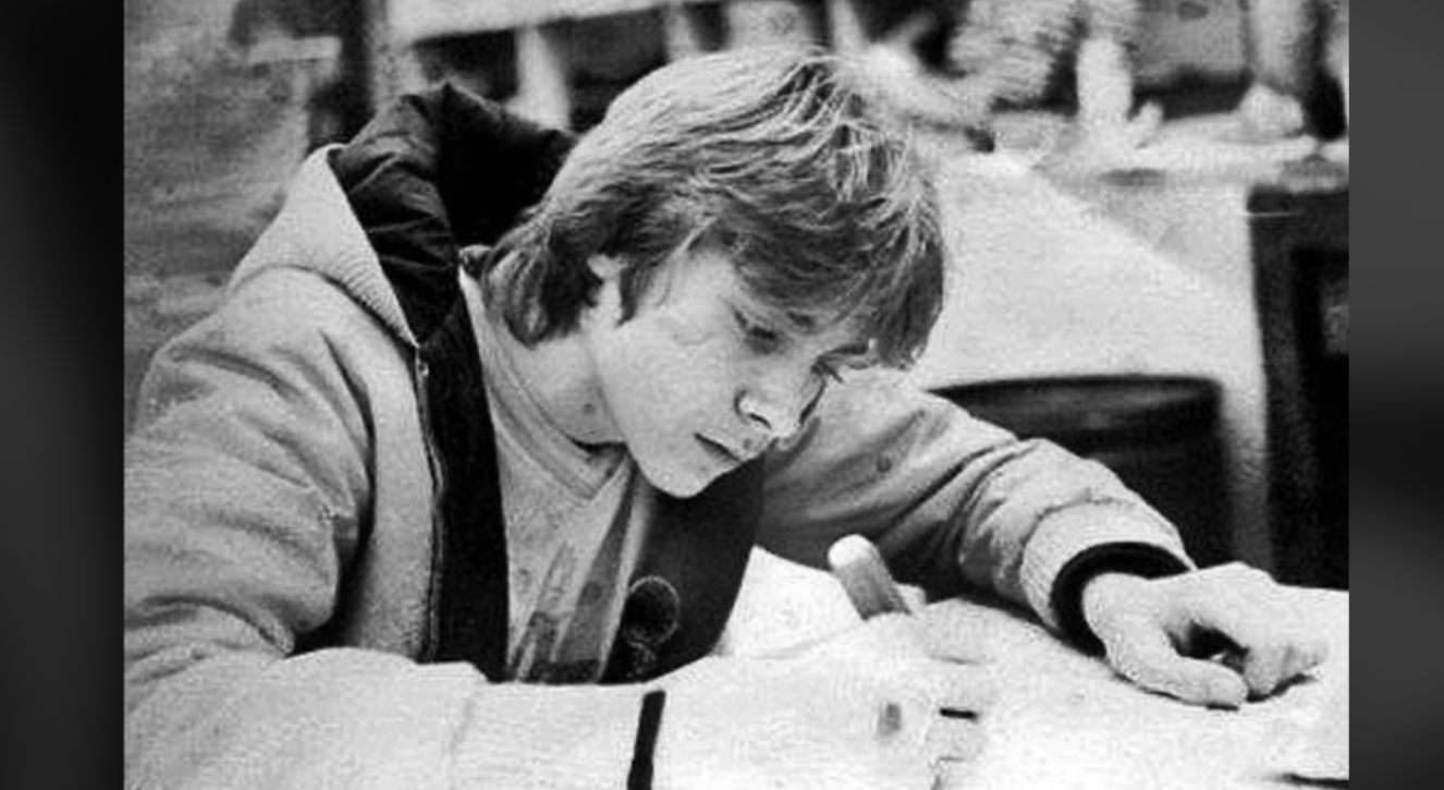 A young person with medium-length hair and a jacket writes intently on paper at a desk in a black-and-white photo, with a blurred background suggesting a classroom or study area.