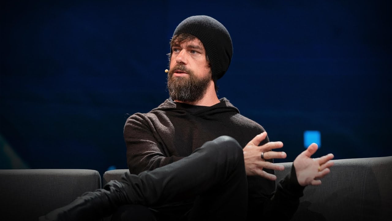 A man with a dark beard, wearing a black beanie and black hoodie, sits on a gray couch with his legs crossed, gesturing with his hands while speaking in front of a dark blue background.