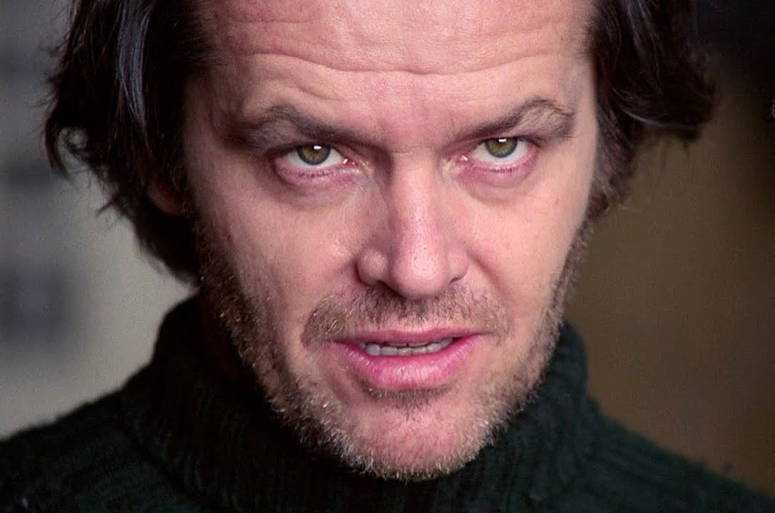 A man with unkempt hair and a scruffy beard stares intensely, wearing a dark sweater. His expression is menacing, with narrowed green eyes and slightly parted lips, suggesting tension or aggression.