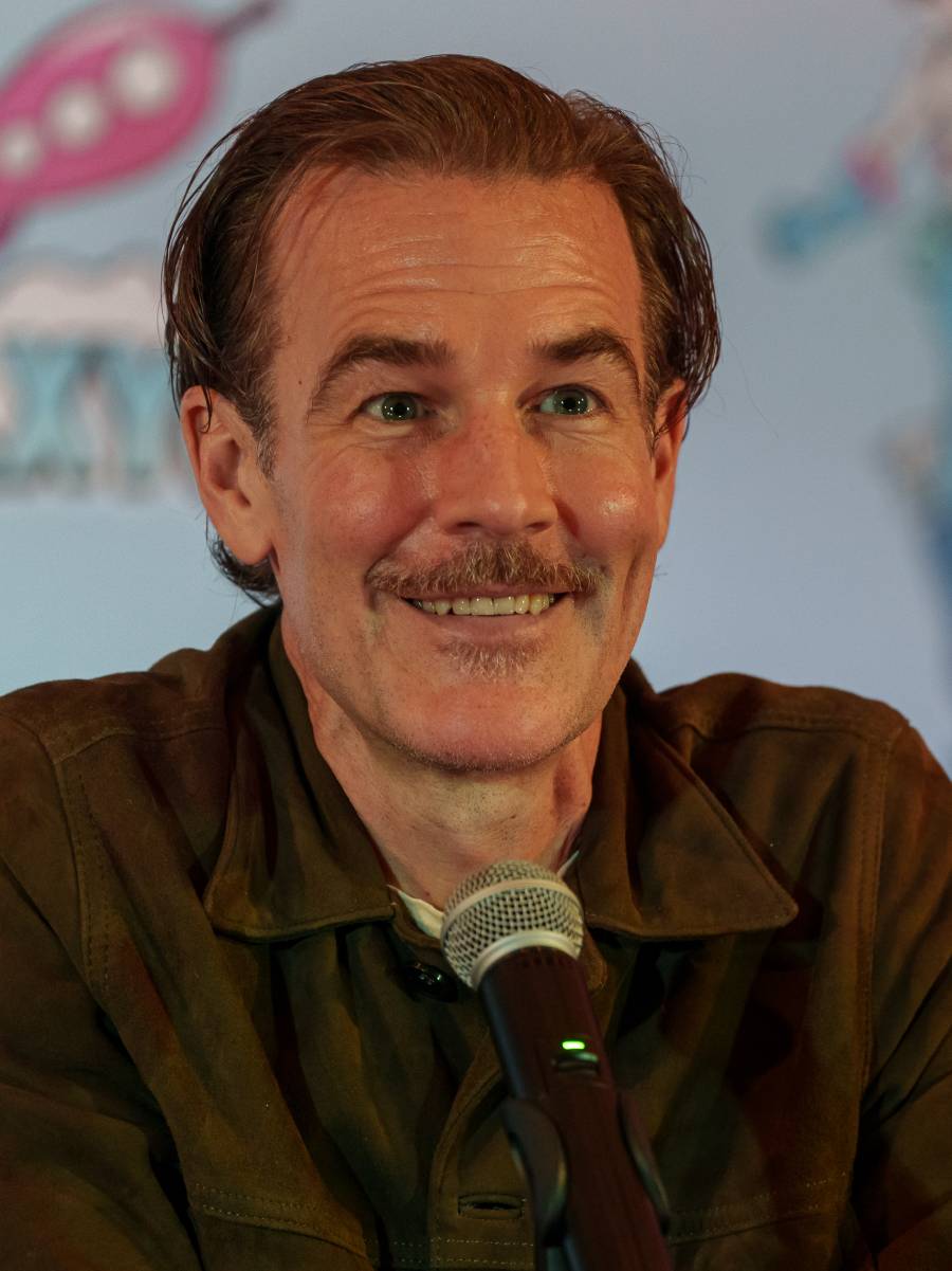 A man with brown hair and a mustache smiles while sitting at a table with a microphone; he is wearing a dark brown jacket and there is a colorful, blurred background behind him.