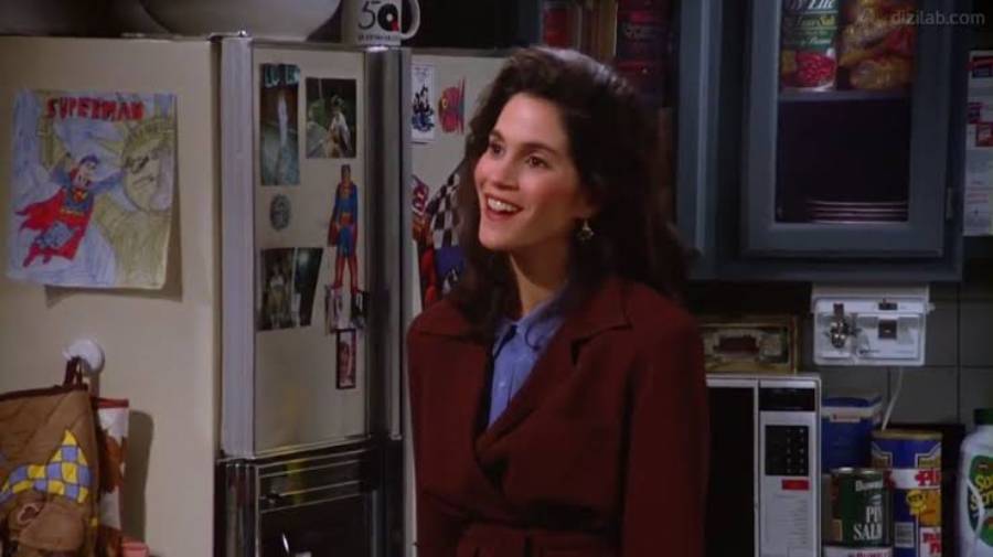 A woman with dark hair, wearing a maroon coat, smiles while standing in a kitchen. Behind her, the fridge is decorated with Superman pictures and magnets, with shelves of food visible to the right.