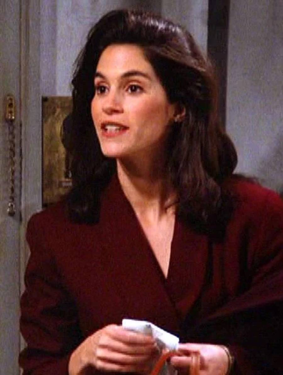 A woman with long dark hair wearing a burgundy blazer stands indoors, holding some items in her hands and speaking. The background features a door and a light-colored wall.