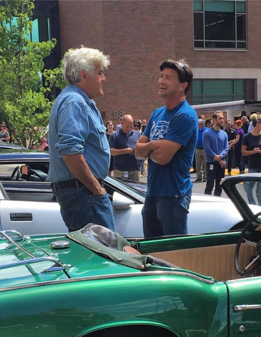 Two men stand and chat by a green classic convertible car at an outdoor event, with a crowd and a brick building in the background. Both appear to be smiling and enjoying the conversation.